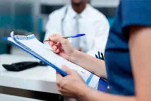 Médico de jaleco atende uma paciente que preenche uma ficha numa prancheta.