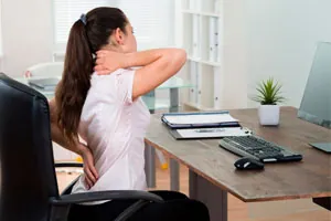 Mulher sentada à mesa do escritório leva a mão ao pescoço dolorido.