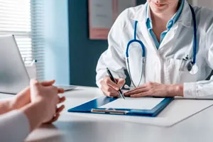 Médico de jaleco faz anotações numa prancheta sobre a mesa do consultório.