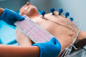 Paciente deitado com eletrodos no peito; a mão de luva segura o traçado do eletrocardiograma.