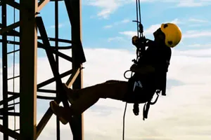 Trabalhador suspenso por cordas numa estrutura metálica, contra o céu.