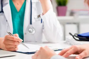 Médica de jaleco e estetoscópio escreve numa prancheta durante a consulta.