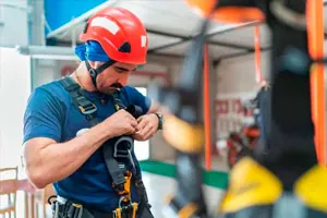 Trabalhador de capacete vermelho ajusta o cinto de segurança tipo paraquedista.