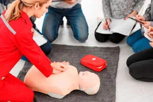 Instrutora demonstra massagem cardíaca num boneco de treinamento, cercada por alunos.