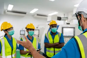 Instrutor orienta trabalhadores de capacete, máscara e colete refletivo.
